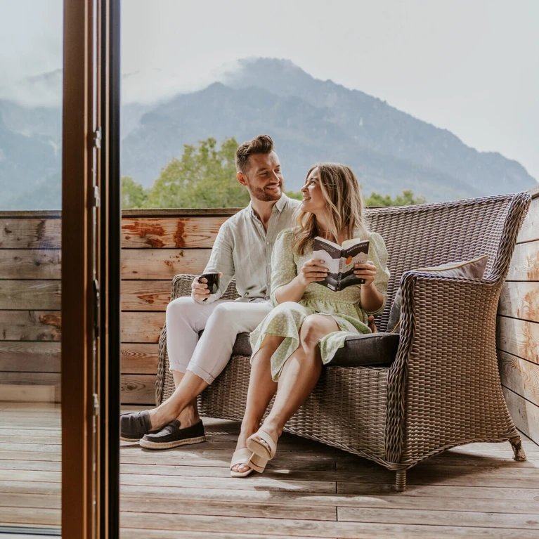 Ein Paar entspannt auf einer Terrasse mit Bergblick am Hotel Klosterhof, sie liest ein Buch, er hält eine Tasse.