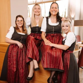 Vier Frauen in traditioneller Tracht lächeln fröhlich in einem modernen Hotelinterieur. Perfekt für das Wellnesshotel Klosterhof in Bayern.