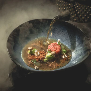 Gourmet dish with broth being poured from a teapot, featuring vibrant ingredients in a dark bowl, served at Hotel Klosterhof.