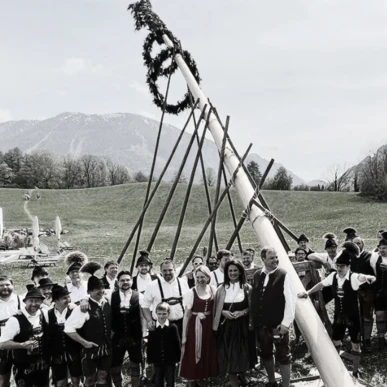 Eine Gruppe in traditioneller Tracht stellt einen Maibaum auf einer Wiese vor dem Hotel Klosterhof auf, im Hintergrund Berge.