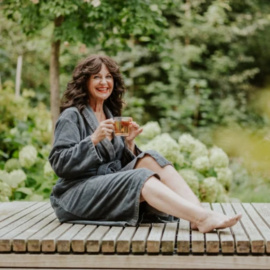 Eine Frau in einem Bademantel sitzt entspannt auf einer Holzterrasse und genießt eine Tasse Tee in einer grünen Gartenlandschaft.