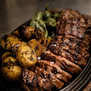 Zarte Steakstücke mit knusprigen Kartoffeln und grünen Peperoni auf einer heißen Platte serviert.
