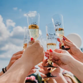 Gläser mit Sekt werden von mehreren Händen in die Höhe gehalten, um auf eine Feier anzustoßen. Heller Himmel im Hintergrund.
