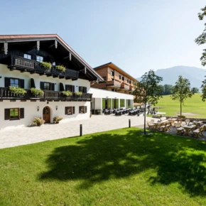 A traditional alpine hotel with balconies and outdoor seating, set against a backdrop of green fields and distant mountains under a clear sky.