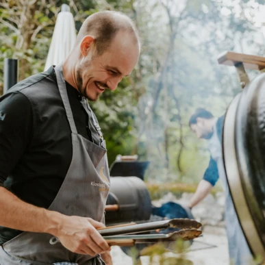 Ein Koch lächelt, während er mit Zangen am Grill in einem Garten arbeitet, umgeben von Bäumen und Pflanzen.