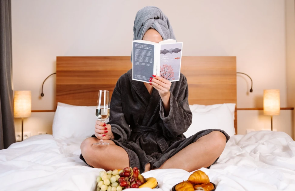 Eine Frau in einem Bademantel sitzt im Bett, liest ein Buch und genießt Sekt, umgeben von frischem Obst und Gebäck im Hotel Klosterhof.