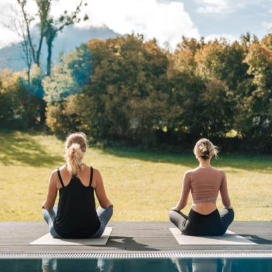 Zwei Frauen meditieren auf Yogamatten im Freien, umgeben von B&auml;umen, mit Blick auf eine gr&uuml;ne Wiese im Hotel Klosterhof, Bayern.