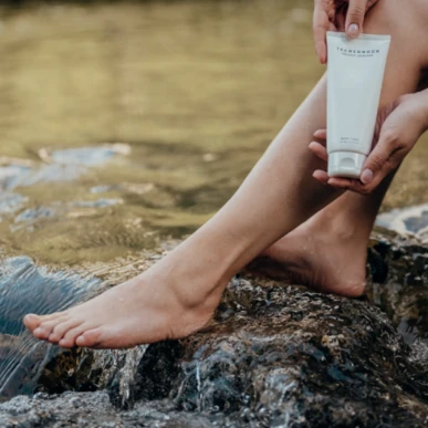 Eine Person hält eine Tube Körperpflegeprodukt am Fuß eines Felsens im Wasser, umgeben von Natur, ideal für Wellness im Hotel Klosterhof.