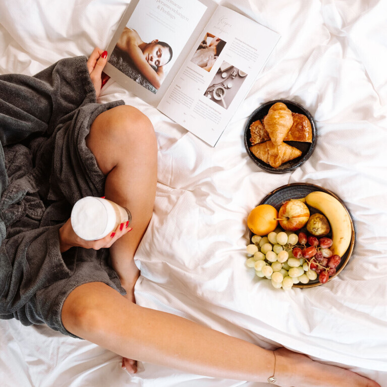 Junge Dame im Bett sitzend mit Magazin, Kaffee, Gebäck und Obstteller.