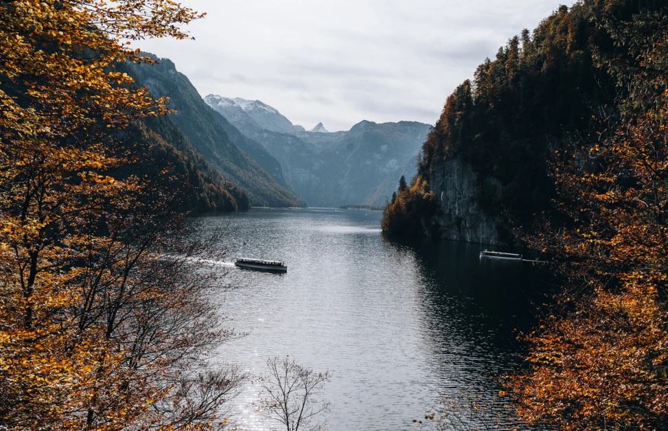 Ein Boot fährt auf dem ruhigen Wasser eines Bergsees, umgeben von herbstlichen Bäumen und majestätischen Bergen im Hintergrund.