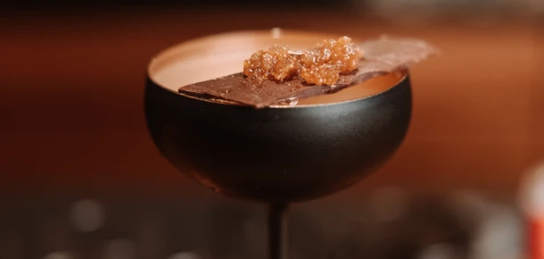 Ein eleganter Cocktail in einer schwarzen Schale, garniert mit Schokolade und Karamell, vor unscharfem Hintergrund.