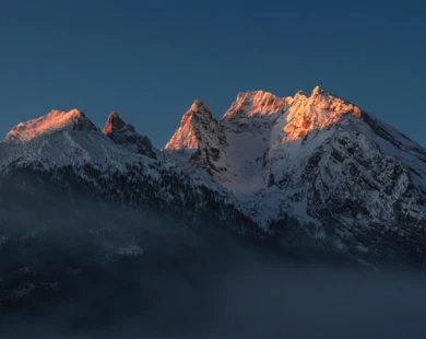 Majestätische Berglandschaft mit schneebedeckten Gipfeln, beleuchtet von der Morgensonne, umgeben von sanften Nebelschwaden.