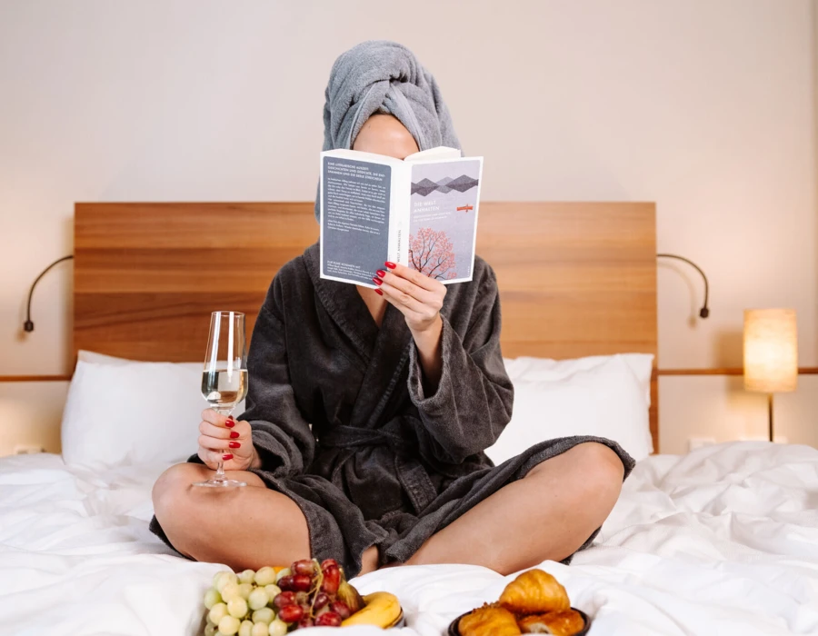 Woman in a robe with a towel on her head, sitting on a bed, reading a book, holding a glass of wine, with fruit and pastries in front.