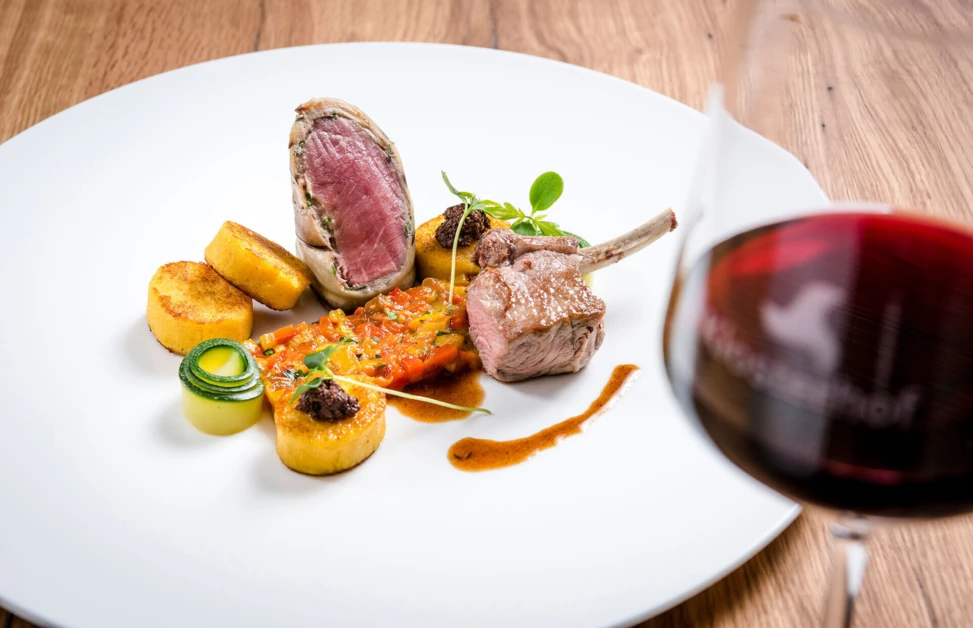 Elegant dish with lamb, vegetables, and sauce, served with a glass of red wine on a wooden table.