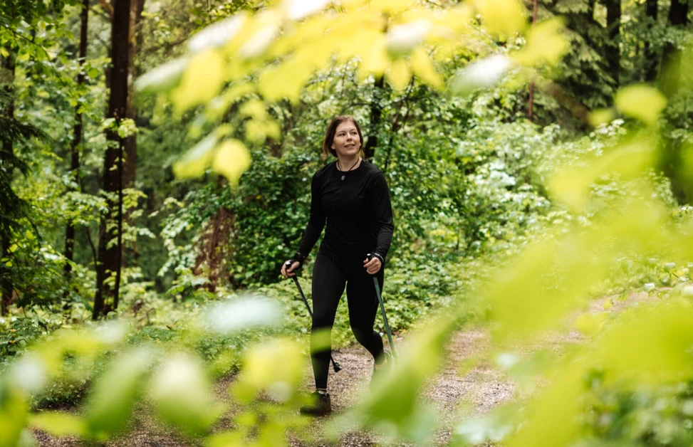 Eine Frau wandert mit Stöcken auf einem Pfad durch einen grünen Wald, umgeben von lebhaften Blättern und Bäumen.