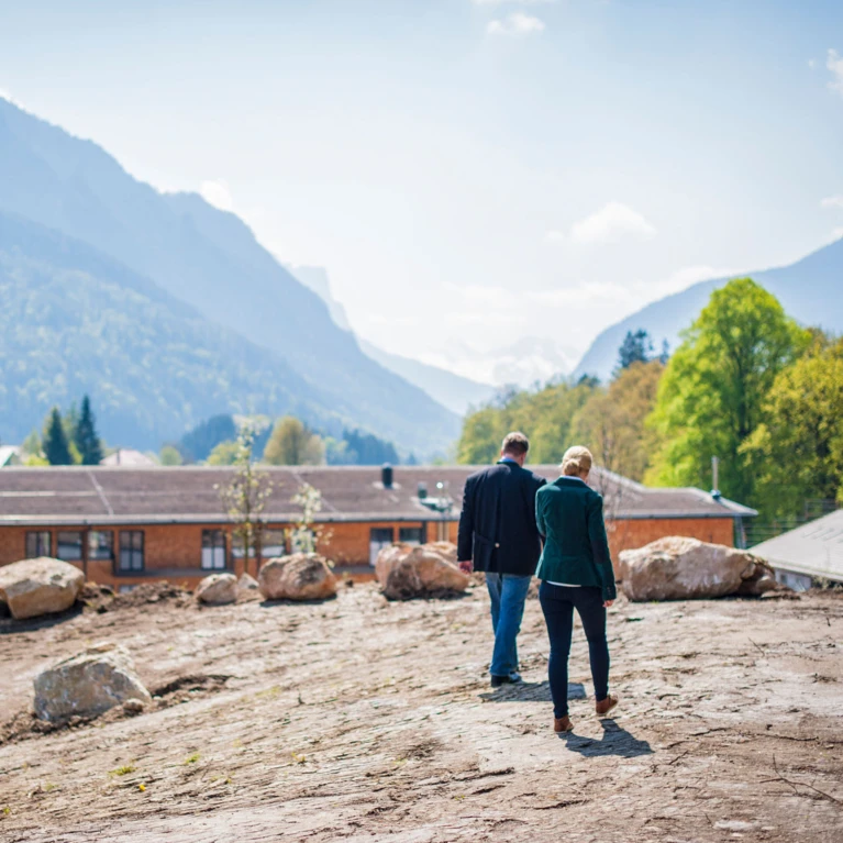 Two people walking on a dirt path with mountains and trees in the background.