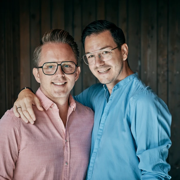 Zwei Männer posieren lächelnd vor einer Holzpaneelwand, einer trägt ein pinkes Hemd und der andere ein blaues.