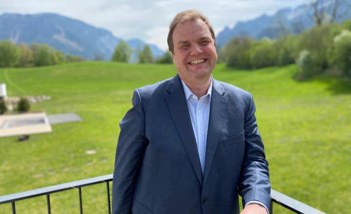 A man in a suit smiles while standing on a balcony with a scenic mountain and green field background.