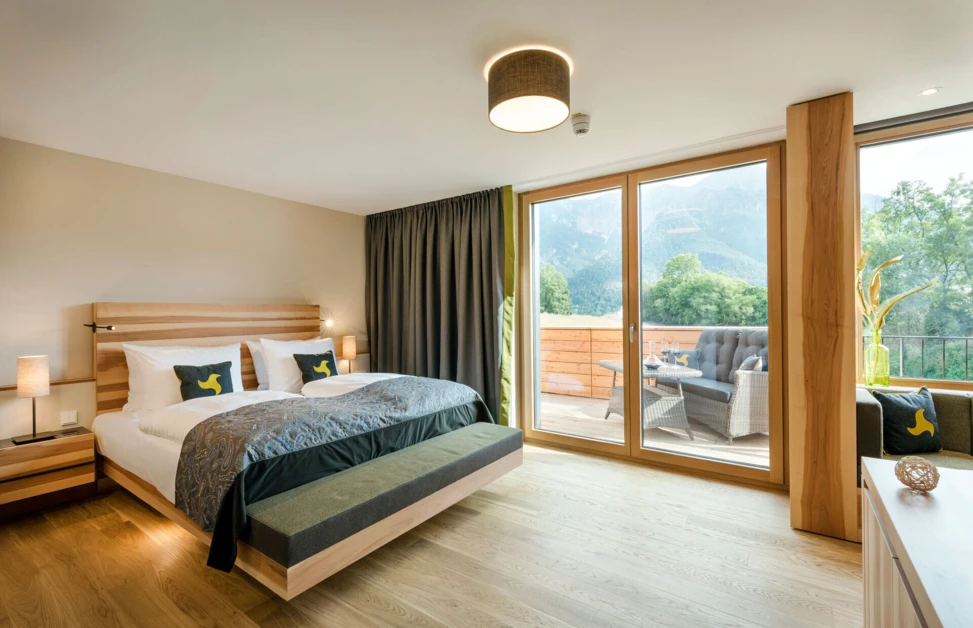 Modernes Hotelzimmer mit großem Bett, Holzboden und großer Fensterfront. Terrasse mit Sitzmöbeln und Blick auf die Berge im Hintergrund.