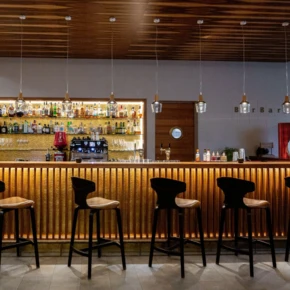Elegante Bar mit Holztheke und stilvollen Hockern, beleuchtet von modernen Pendelleuchten, hinter der eine Vielzahl von Getränken präsentiert wird.