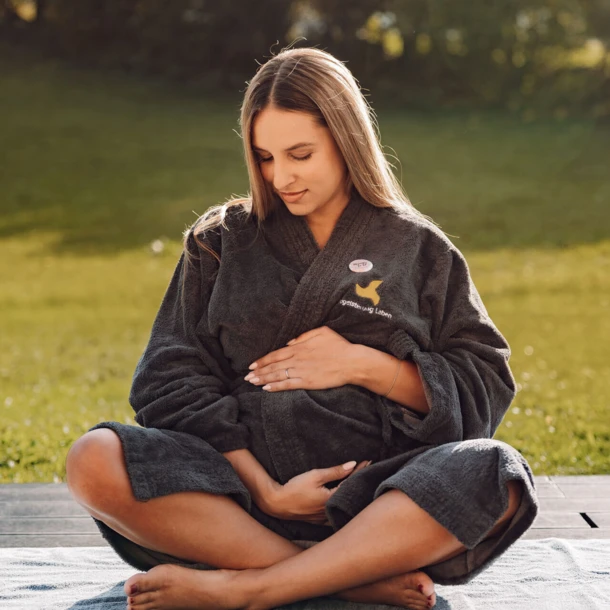 Frau in einem grauen Bademantel sitzt im Freien und hält sanft ihren Bauch. Hintergrund zeigt eine grüne Wiese im Wellnesshotel Klosterhof.