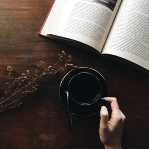 An open book, a hand holding a cup of coffee, and dried flowers on a wooden table.