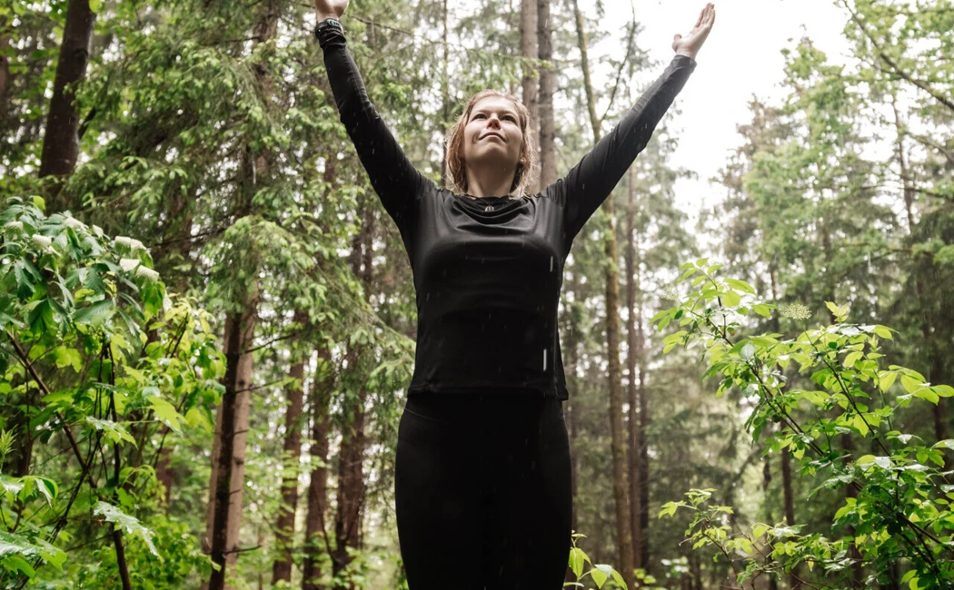 Eine Person steht in einem Wald mit erhobenen Händen, umgeben von grünen Pflanzen und hohen Bäumen, und genießt die Natur.