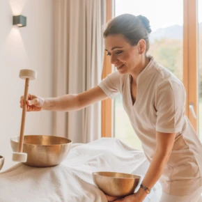 Eine Therapeutin nutzt Klangschalen zur Entspannung in einem hellen, ruhigen Raum mit gro&szlig;en Fenstern und Blick ins Gr&uuml;ne.