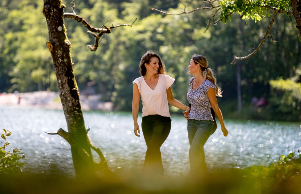 Two women walking and smiling by a tranquil lake surrounded by lush greenery at Wellnesshotel Klosterhof.