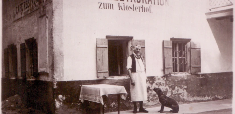 Ein Mann in Schürze steht vor dem Café Restauration zum Klosterhof, neben einem Hund und einem Tisch. Historische Szenerie in Bayern.