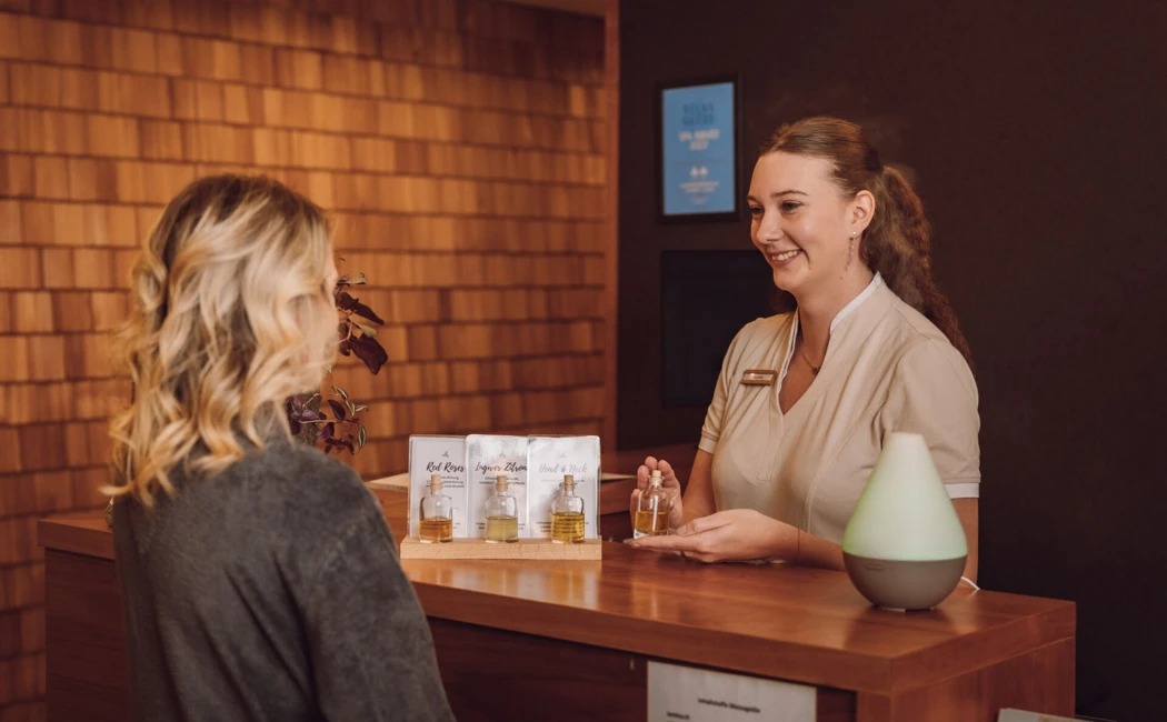 Eine Mitarbeiterin berät eine Kundin am Empfang, präsentiert Pflegeprodukte und einen Aromadiffuser in einem stilvollen Ambiente.