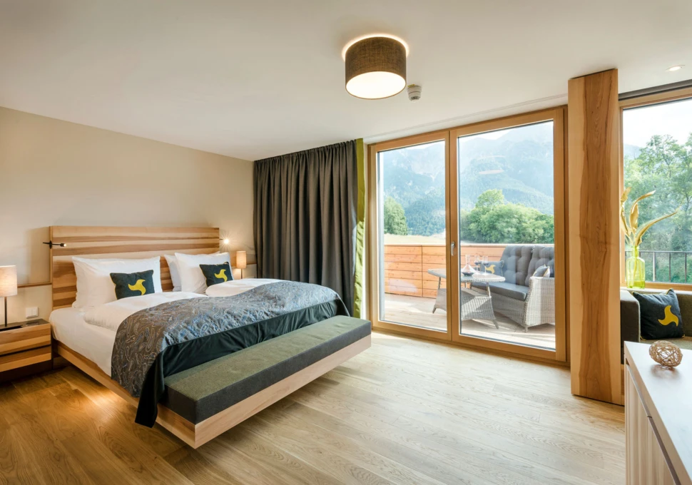 Modernes Hotelzimmer mit gro&szlig;em Bett, Holzboden und gro&szlig;er Fensterfront. Terrasse mit Sitzm&ouml;beln und Blick auf die Berge im Hintergrund.