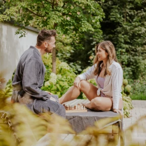 Ein Paar sitzt in Freizeitkleidung im Garten und spielt Schach auf einem Holztisch. Sie lächeln und genießen die entspannte Atmosphäre.