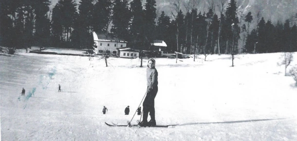 Eine Skifahrerin steht auf einer verschneiten Piste mit Bergen im Hintergrund, umgeben von Bäumen.