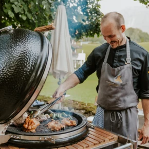 Ein Koch grillt Fleisch und Gemüse auf einem großen Grill im Freien, umgeben von einer schönen, grünen Landschaft.