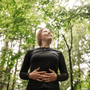 Eine Frau in einem Wald lächelt und hält ihre Hände auf ihrem Bauch, umgeben von grünen Bäumen. Wellness-Atmosphäre im Hotel Klosterhof.
