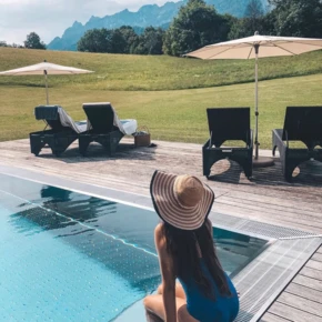 Woman in blue swimsuit and sun hat sitting by a pool, overlooking green fields and mountains at a serene resort.