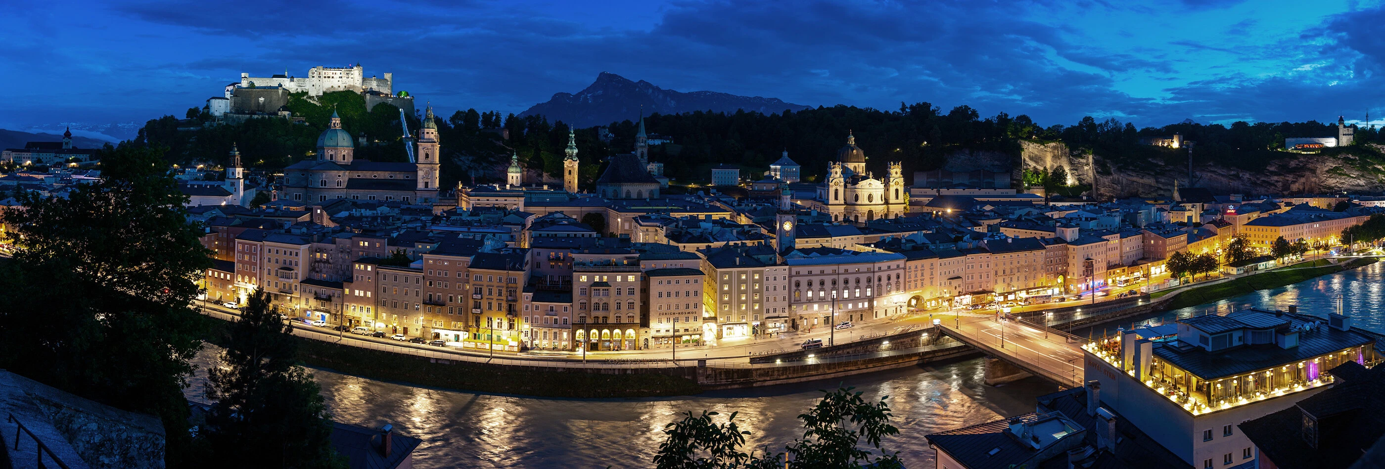 Panoramaansicht von Salzburg bei Nacht, mit beleuchteten Gebäuden und der Festung im Hintergrund. Einladende Atmosphäre am Fluss.