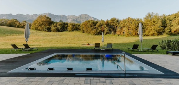 Ein moderner Pool umgeben von Liegen und Sonnenschirmen, mit Blick auf eine gr&uuml;ne Wiese und die Berge im Hintergrund.