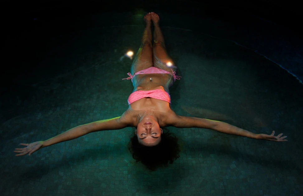 Woman floating calmly in a dimly lit pool, wearing a pink bikini, arms spread wide.