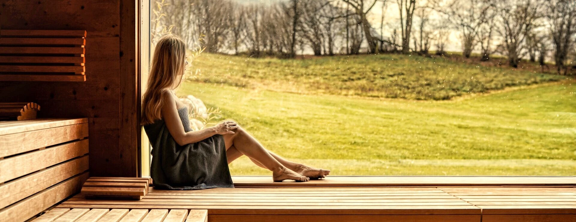 Eine entspannte Frau sitzt in einem modernen Sauna-Bereich und blickt auf die grüne Landschaft durch große Fenster.
