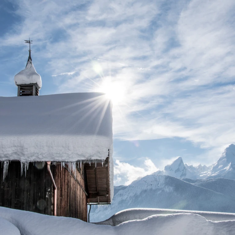 Schneebedecktes Dach eines Hauses mit Sonnenstrahlen und Bergen im Hintergrund, unter blauem Himmel mit Wolken.