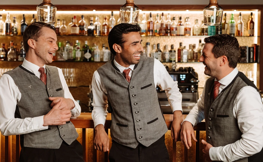 Drei lächelnde Barkeeper in eleganter Kleidung stehen an einer Bar, umgeben von Spirituosenflaschen im Hotel Klosterhof.