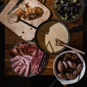 Ein rustikaler Tisch mit Käsefondue, frischem Brot, Wurstplatten und Kartoffeln, umgeben von frischem Salat.