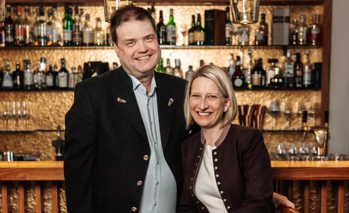 Ein lächelndes Paar posiert vor der Bar im Hotel Klosterhof, umgeben von Spirituosen. Das Wellnesshotel bietet eine gemütliche Atmosphäre.