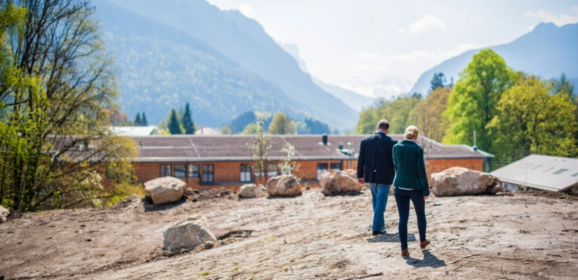 Die Inhaber, Henrike und Andreas Färber, spazieren über die Baustelle, mit Blick auf die umliegende Bergwelt.