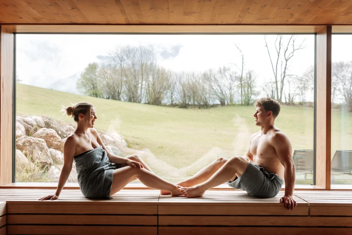 Paar entspannt in einer modernen Sauna mit Blick auf die grüne Landschaft. Natürliche Lichtverhältnisse schaffen eine ruhige Atmosphäre.