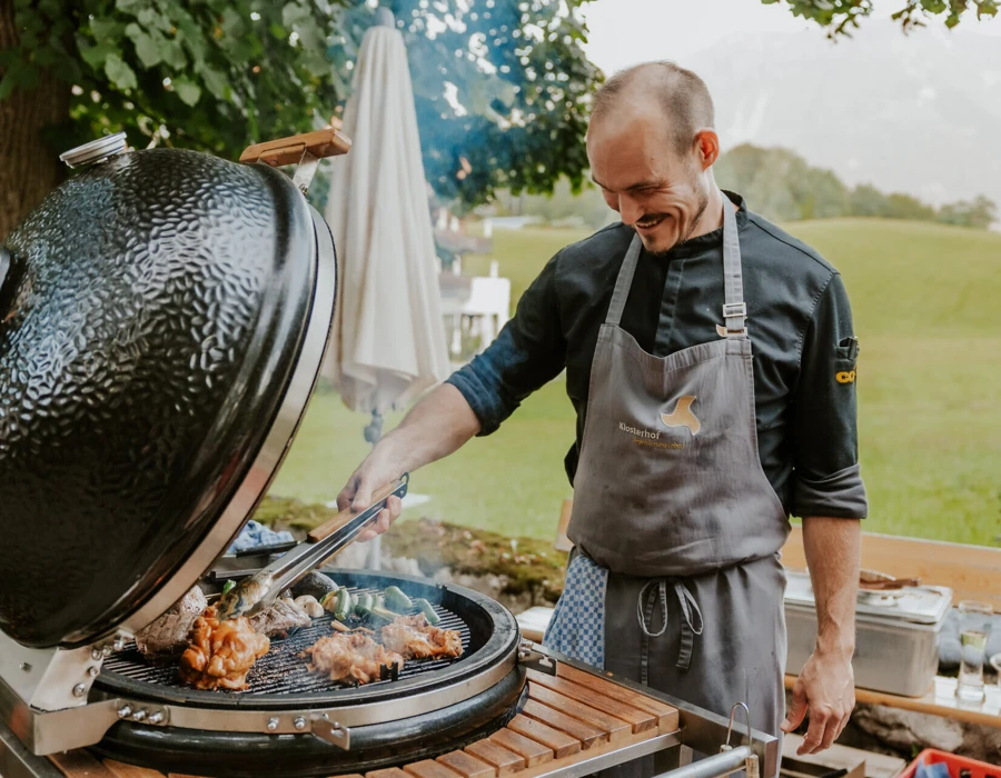 Ein Koch grillt Fleisch und Gem&uuml;se auf einem gro&szlig;en Grill im Freien, umgeben von einer sch&ouml;nen, gr&uuml;nen Landschaft.