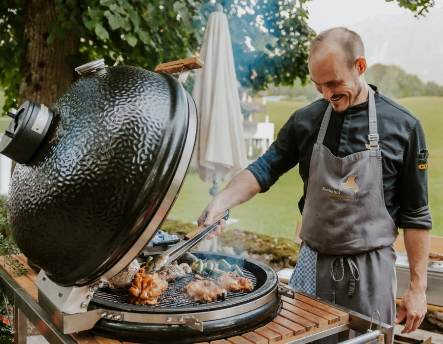 Ein Koch grillt Fleisch und Gem&uuml;se auf einem gro&szlig;en Grill im Freien, umgeben von einer sch&ouml;nen, gr&uuml;nen Landschaft.
