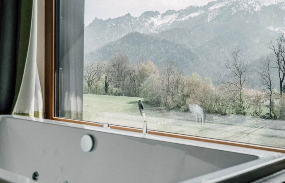 Bathtub with a view of snow-capped mountains and green fields through a large window, creating a serene and relaxing atmosphere.